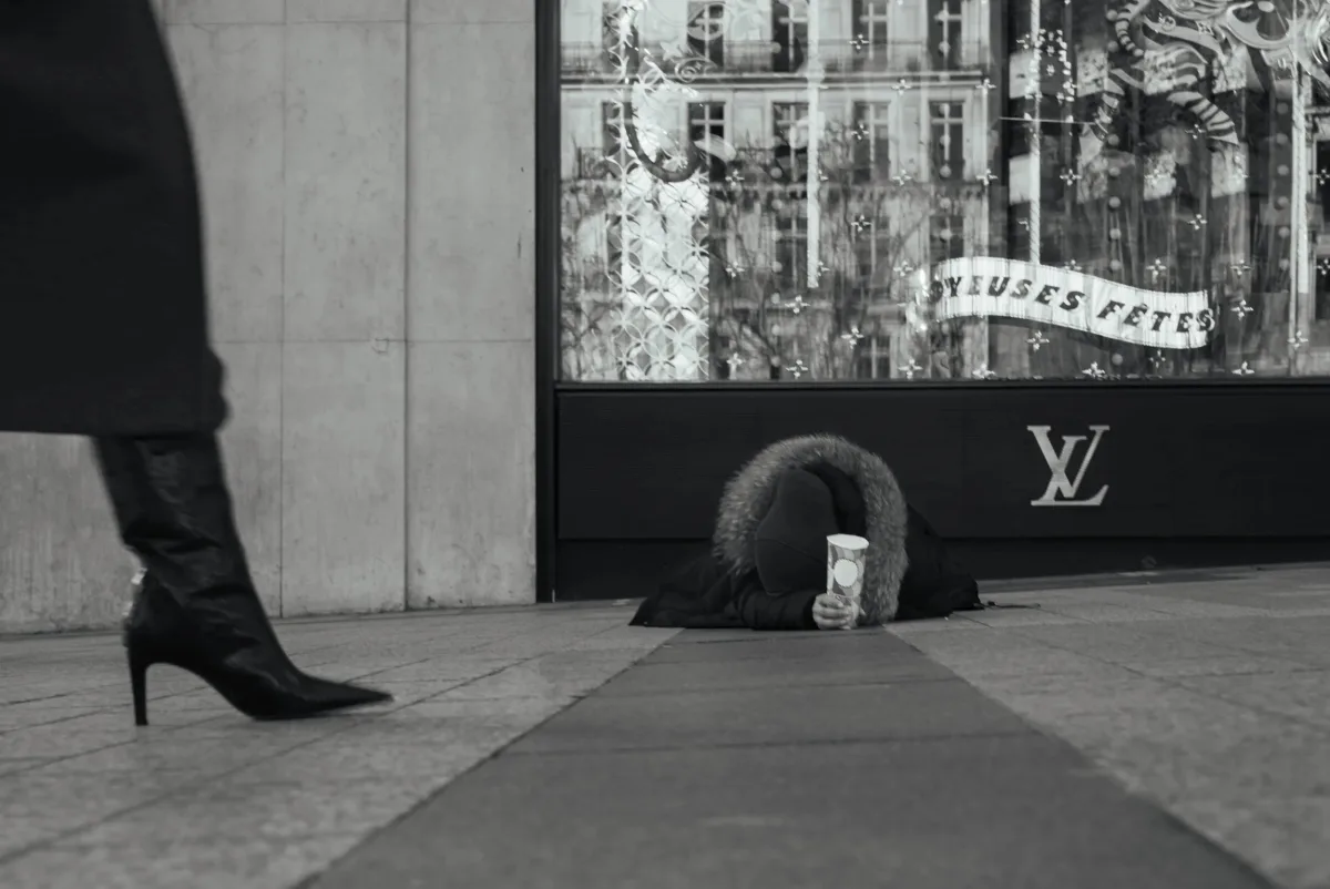 A person lying on the sidewalk with a coat with a fur-lined hood, holding a cup. A woman walking past is visible, with only her legs and black high-heeled boots in the frame. The background shows a shop window with reflections and the Louis Vuitton logo.