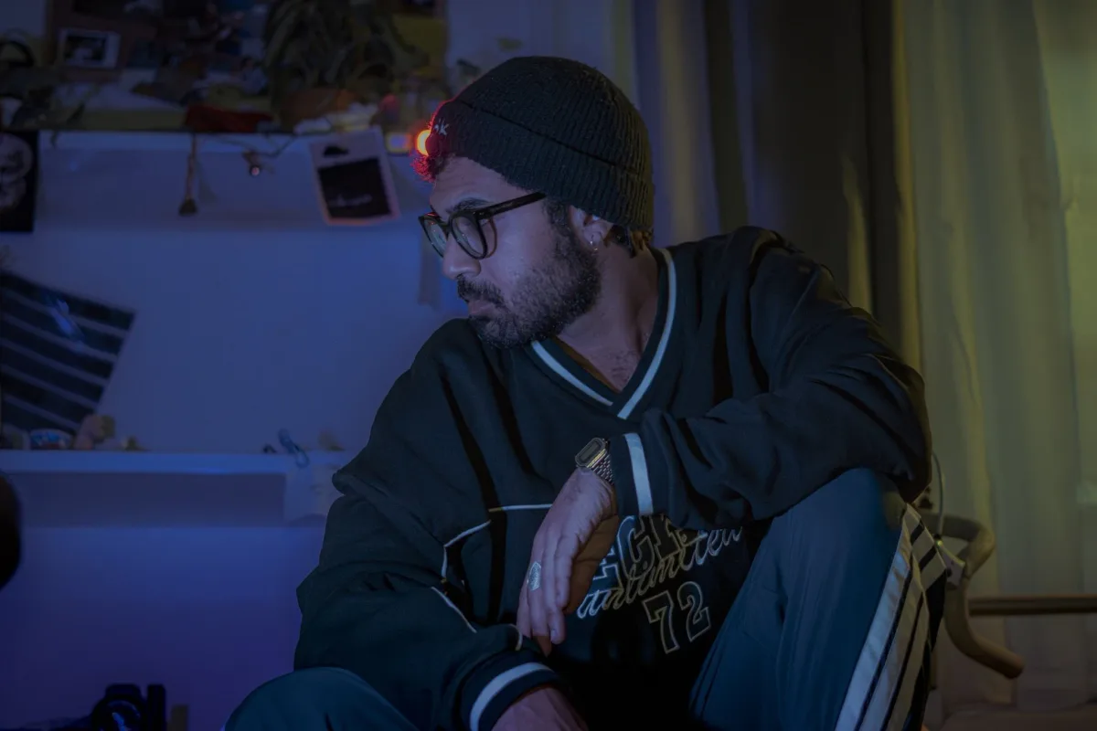A man with glasses, a beard, and a beanie, sitting and looking down in a dimly lit room with yellow curtains behind him. Parham Motamedifard